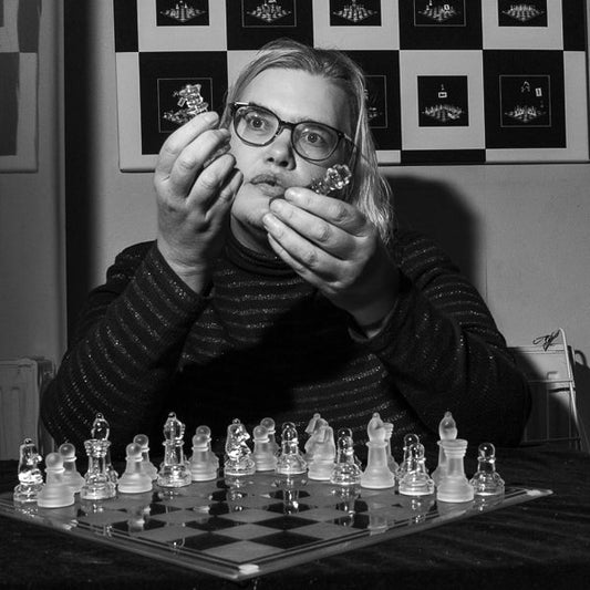 Square black and white photograph of Nancy Pinkerton Looking at the two chess pieces she is holding Above a chess board.