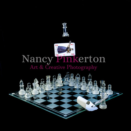 Photographic art print with image of a playing card featuring Mary Queen of Scots and an axe floating above a glass chessboard with chess pieces laid out in rows. A Queen piece is at the front with a quill signing a piece of paper.