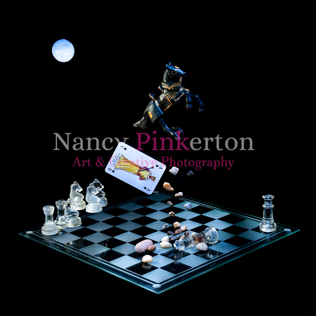 Photographic art print with image of a playing card featuring King Alexander the III and small rocks falling from a model horse floating above a glass chessboard with the king piece lying on the ground and other chess pieces strategically placed.