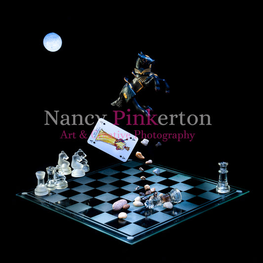 Photographic art print with image of a playing card featuring King Alexander the III and small rocks falling from a model horse floating above a glass chessboard with the king piece lying on the ground and other chess pieces strategically placed.